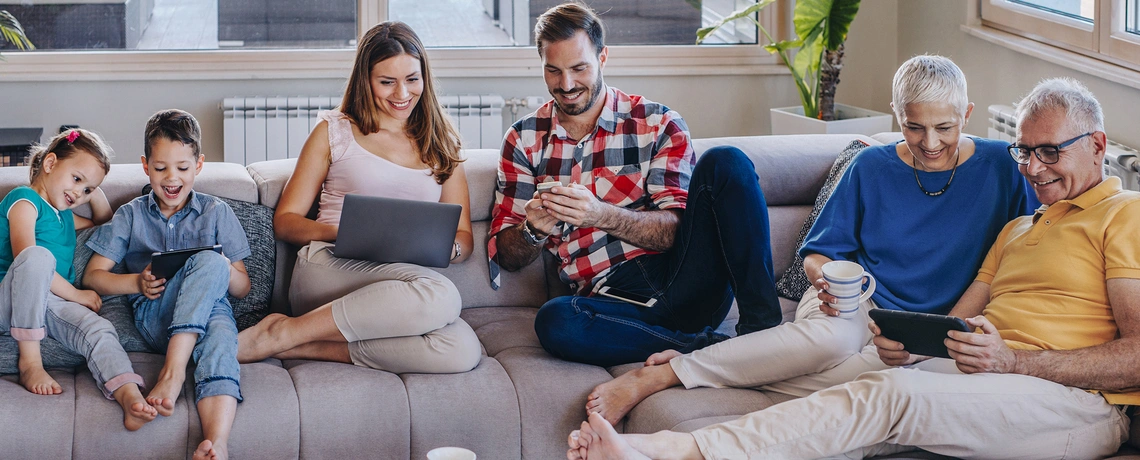 Eine Familie sitzt auf dem Sofa und alle lächeln. Einige Personen halten ein Tablet oder Laptop in der Hand und lesen die digtiale Zeitung.
