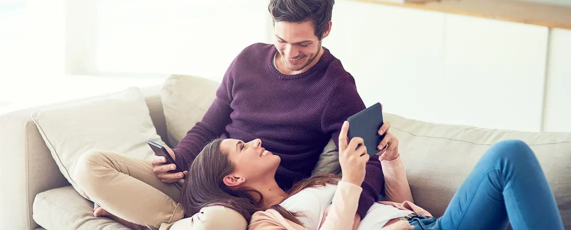 Eine Frau und ein Mann liegen auf dem Sofa. Er hat ein Smartphone in der Hand, sie ein Tablet.